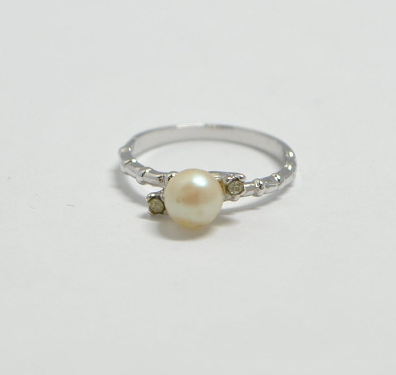 May include: A silver ring with a white pearl and two small clear stones. The band of the ring is textured.