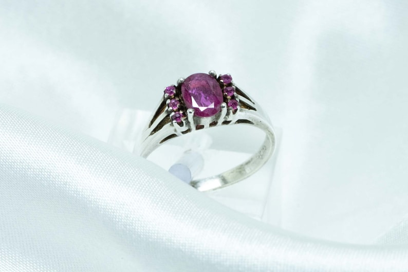 Peut inclure: Bague en argent ornée d'un rubis rose foncé ovale au centre, entouré de petits rubis assortis. La bague présente un design à tige fendue. Le bijou est présenté sur un fond de satin blanc.