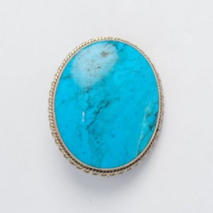 May include: A silver oval brooch with a large turquoise stone set in the center. The stone has a blue and green color with white veins.