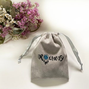 May include: A grey velvet pouch with a drawstring closure. The pouch has the text "IROQUOISE" printed on it in black with a stylised dreamcatcher design.