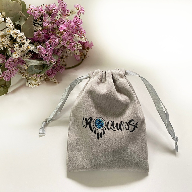 May include: A grey velvet pouch with a drawstring closure. The pouch has the text "IROQUOISE" printed on it in black with a stylised dreamcatcher design.