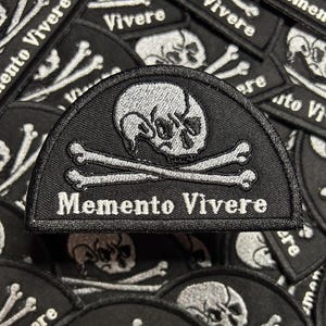 May include: Black and white embroidered patch featuring a skull and crossbones design with the words "Memento Vivere". The patch has a semi-circular shape and is surrounded by other similar patches.