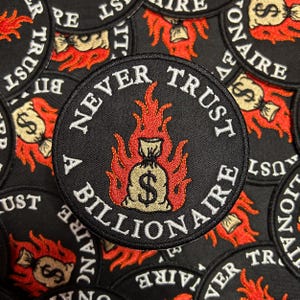 Puede incluir: Un parche bordado en blanco y negro con el texto "Never Trust A Billionaire" en un círculo. Una bolsa de dinero está en llamas en el centro del parche.