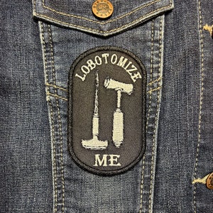 May include: A black and white embroidered patch on a blue denim jacket. The patch says "lobotomize me" with a hammer and a scalpel.