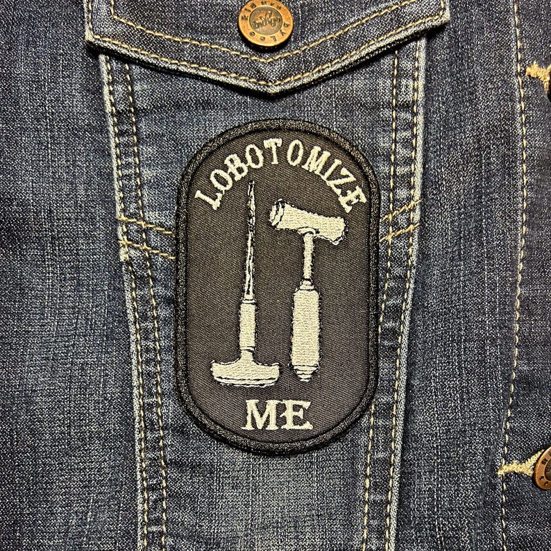 May include: A black and white embroidered patch on a blue denim jacket. The patch says "lobotomize me" with a hammer and a scalpel.