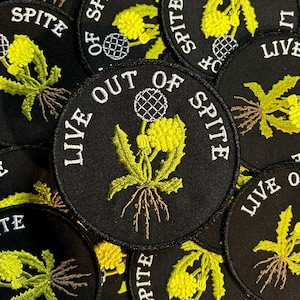 May include: Black embroidered patches with the phrase "LIVE OUT OF SPITE" in white, surrounding a yellow and green plant design. The patches are round and feature the word "SPITE" in white around the edge.