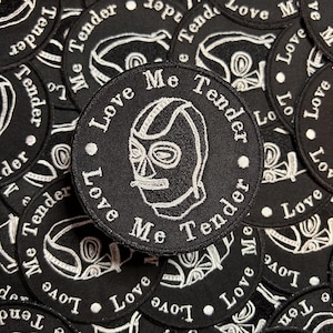 May include: A close-up of a stack of black embroidered patches with a white wrestler wearing a mask. The text on the patches reads "Love Me Tender".
