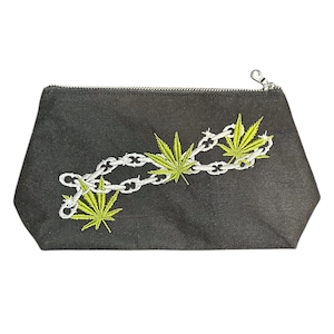 May include: Black zippered pouch with an embroidered design featuring a chain and cannabis leaves in white and green. The pouch has a silver zipper and a small loop for attaching to a keychain.