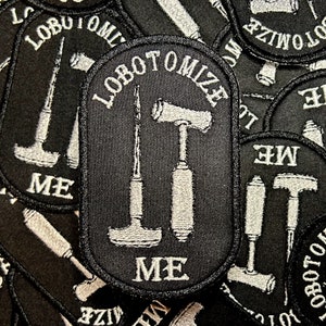 May include: Close-up of a black embroidered patch with white stitching. The patch features three tools: a hammer, a chisel, and a screwdriver. The text "lobotomize me" is written in white on the patch.