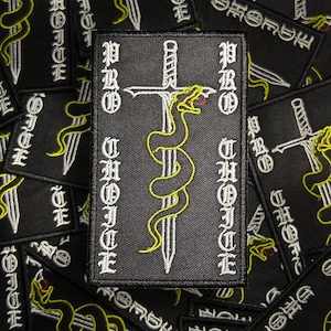 May include: Black embroidered patch with a white sword and a yellow snake wrapped around it. The text "PROHIBIT" is written vertically on both sides of the sword.