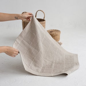 May include: A waffle weave linen bath towel in a natural beige color. The towel is folded and being held by a hand.