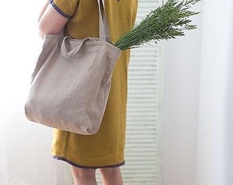 May include: A person wearing a mustard yellow dress with a light gray tote bag. The bag is made of linen and has a long strap. The person is holding a bouquet of green flowers in the bag.