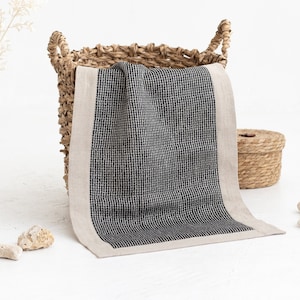 Puede incluir: Una toalla a cuadros blancos y negros con un borde beige está doblada en una cesta de mimbre. El patrón de la toalla es una cuadrícula de pequeños cuadrados. Una pequeña cesta redonda de mimbre está en el fondo.