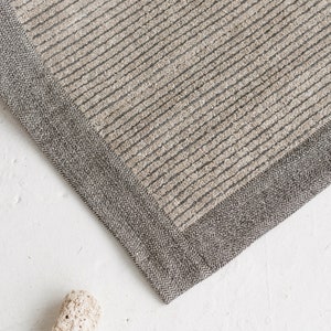 May include: A close-up of a beige and gray striped rug with a herringbone border. The rug is made of a soft, textured material.