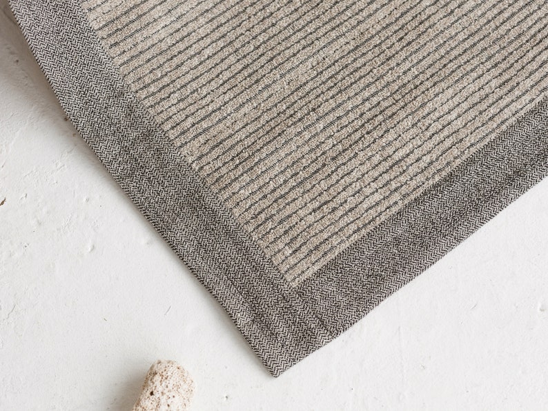May include: A close-up of a beige and gray striped rug with a herringbone border. The rug is made of a soft, textured material.