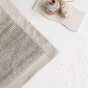 May include: A beige bath mat with a textured, striped pattern and a solid beige border. The mat is on a white surface.