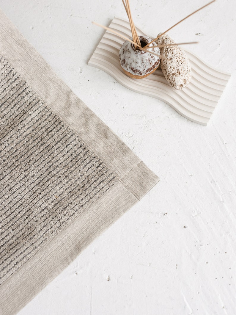 May include: A beige bath mat with a textured, striped pattern and a solid beige border. The mat is on a white surface.