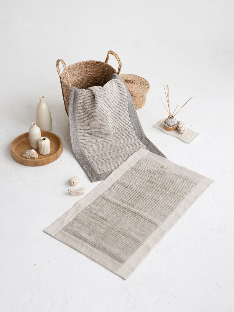 May include: A set of two bath mats in a natural linen color with a gray stripe pattern. The mats have a white border and are made of a soft, absorbent material. The mats are displayed on a white surface with a wicker basket, vases, and other decorative items.