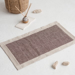 Op de afbeelding: Een rechthoekig tapijt met een geweven, donkerrood midden en een lichtbeige rand. Het tapijt ligt op een wit oppervlak, met een geweven mand, een keramische diffuser en decoratieve stenen op de achtergrond. Het tapijt meet ongeveer 60 x 90 cm.