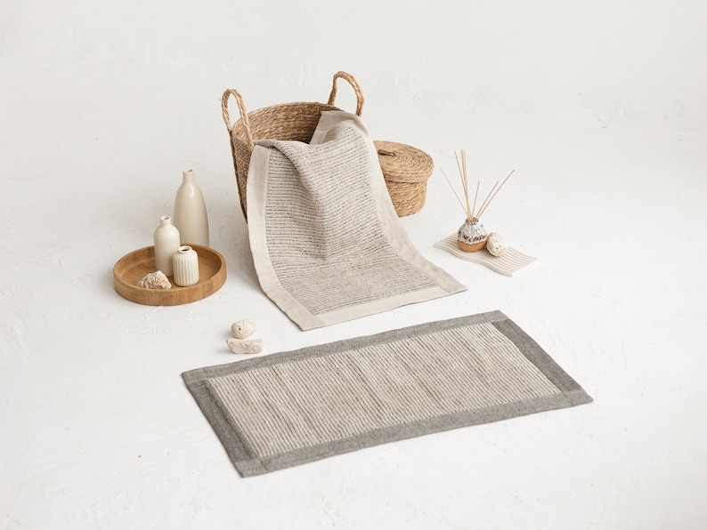 May include: A set of two bath mats in a light gray color with a textured woven design. The mats have a darker gray border. The mats are displayed on a white surface with a wicker basket, vases, and other decorative items.