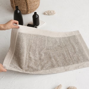 May include: A beige bath mat with a textured surface and a white border. The mat is made of linen and is folded in half.