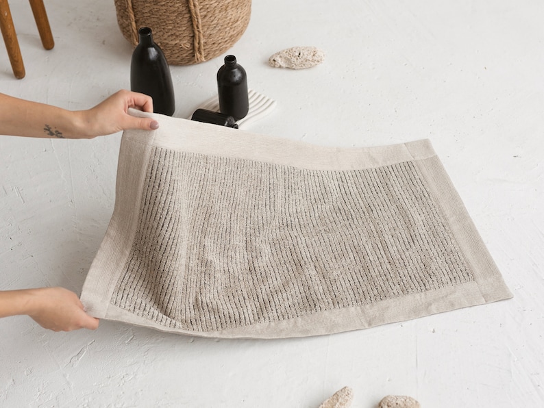 May include: A beige bath mat with a textured surface and a white border. The mat is made of linen and is folded in half.