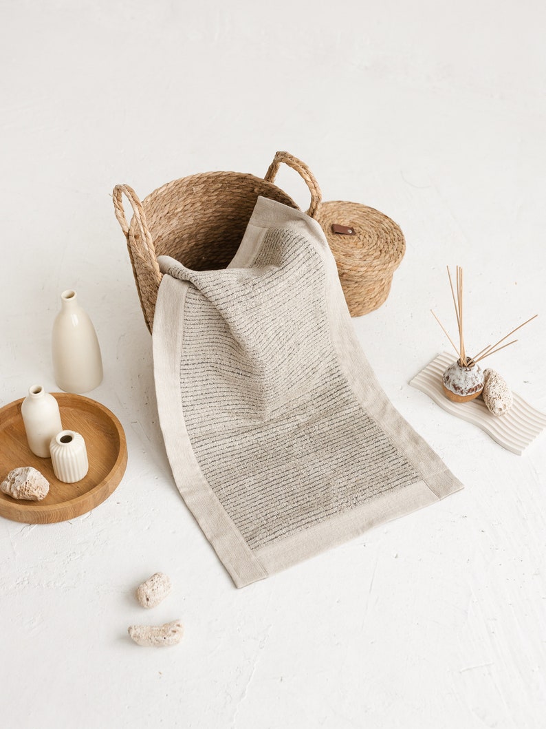 May include: A woven basket with a lid, a white bath mat, and two small white ceramic vases on a wooden tray. The items are arranged on a white surface.