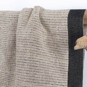 May include: A beige bath towel with black stripes and a black border. The towel is hanging on a wooden branch.
