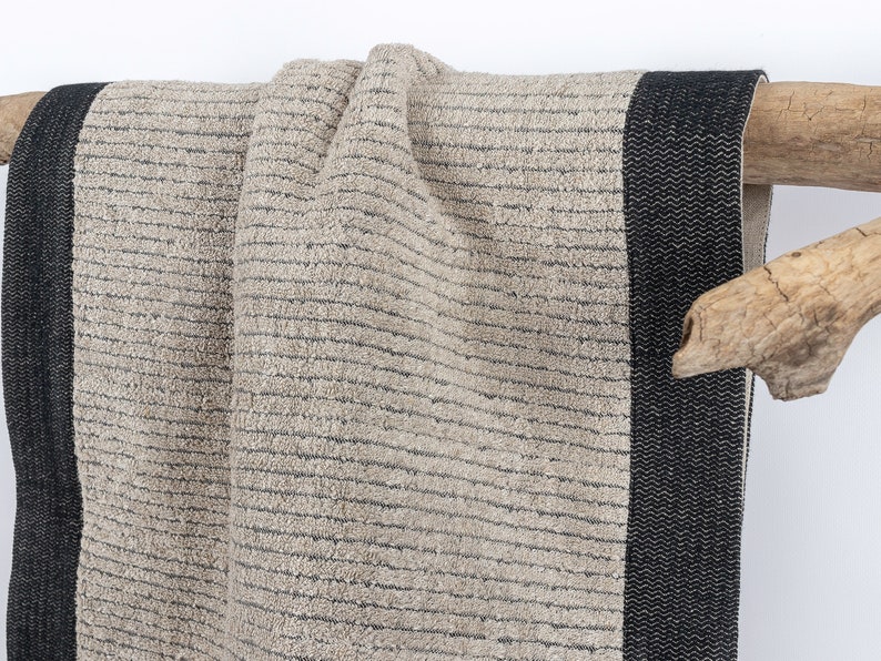 May include: A beige bath towel with black stripes and a black border. The towel is hanging on a wooden branch.