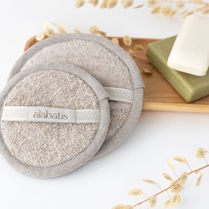 Puede incluir: Dos esponjas de baño redondas, beige y texturizadas con una etiqueta blanca que dice "alabatis". Las esponjas están sobre una superficie blanca con una bandeja de madera que contiene dos barras de jabón en el fondo.