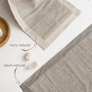 May include: Two bath mats, one ivory with a natural linen border and one black with a natural linen border. Both mats have a ribbed texture.