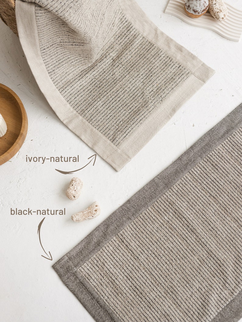 May include: Two bath mats, one ivory with a natural linen border and one black with a natural linen border. Both mats have a ribbed texture.