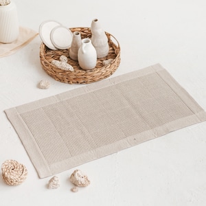 May include: A beige bath mat with a woven texture and a white border. The mat is lying on a white surface with a wicker basket and ceramic vases.