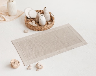 May include: A beige bath mat with a woven texture and a white border. The mat is lying on a white surface with a wicker basket and ceramic vases.