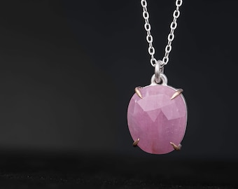 May include: A silver necklace with a large, oval-shaped pink gemstone pendant. The pendant is faceted and held in place by four gold-colored prongs. The chain is a delicate silver link style, and the background is black.