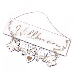 Op de afbeelding: Een wit houten bord met het woord "Willkommen" in cursief geschreven. Het bord heeft een touwtje om het op te hangen. Onder het bord bevindt zich een decoratieve krans van bladeren, vogels en een hart.