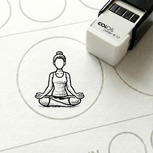 Peut inclure: Un tampon noir et blanc représentant une personne en position de yoga assise, à l'intérieur d'un cercle gris. Le tampon est à côté d'un tampon blanc et noir "COLOR printer Q 12".