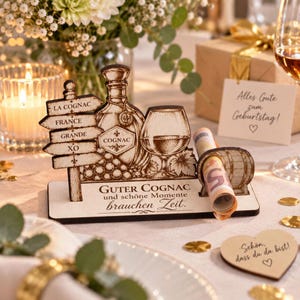Op de afbeelding: Een decoratieve houten standaard met lasergegraveerde illustraties van een cognacfles, een glas en een wegwijzer met de tekst LA COGNAC, FRANCE, GRANDE, XO. De basis bevat de tekst GUTER COGNAC und schöne Momente brauchen Zeit.