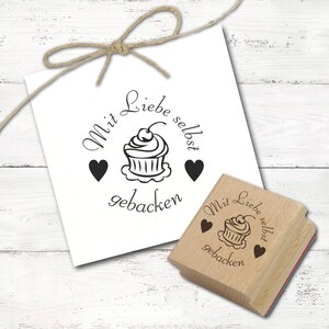 May include: A white card with a black stamp of a cupcake with a cherry on top, two hearts, and the words "Mit Liebe selbst gebacken". A wooden stamp with the same design is also present.