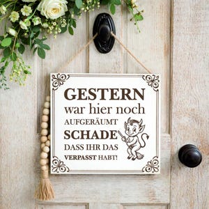 Può includere: Un cartello rettangolare bianco con testo marrone e illustrazione di un diavolo. Il cartello recita "Gestern war hier noch aufgeräumt. Schade dass ihr das verpasst habt!" Elementi floreali decorativi e una ghirlanda di perline di legno visibili.