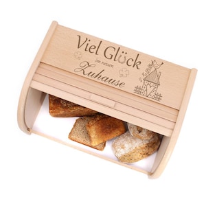 Könnte beinhalten: Eine Brotbox aus Holz mit einem Schiebedeckel. Der Deckel ist mit dem Text "Viel Glück im neuen Zuhause" und einem kleinen Haus mit einem Herzen und einem Vogel graviert. Die Brotbox ist mit Brot gefüllt.