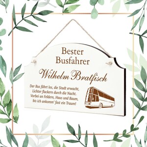 Puede incluir: Un letrero de madera blanco con texto marrón que dice "Bester Busfahrer Wilhelm Bratfisch". El letrero tiene un colgador de cuerda y una pequeña ilustración de un autobús con el texto "Der Bus fährt los, die Stadt erwacht, Lichter flackern durch die Nacht. Vorbei an Feldern, Haus und Baum, bis ich ankomm' fast ein Traum!"