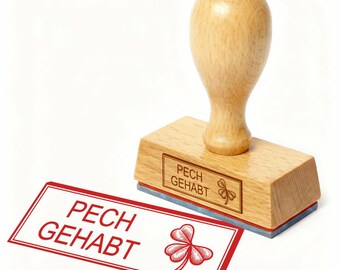 Può includere: Un timbro in legno con le parole "PECH GEHABT" e un disegno a trifoglio. Il timbro è accanto a un rettangolo bordato di rosso con lo stesso testo e trifoglio. Il timbro ha un manico in legno.