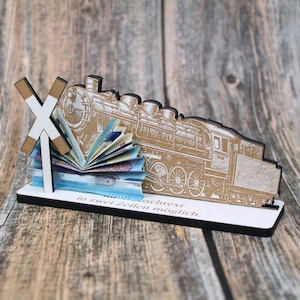 May include: A wooden train-shaped display with an engraved design. It holds a fan of colourful paper money and a miniature railroad crossing sign. The base has text in German.