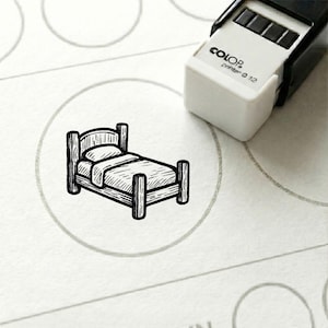 Op de afbeelding: Een zwart-wit stempel van een bed in een cirkel. De stempel staat op een wit oppervlak, met een 'Color printer Q 12' stempel in de rechterbovenhoek. Het bed heeft een hoofdbord, voetbord en een deken.