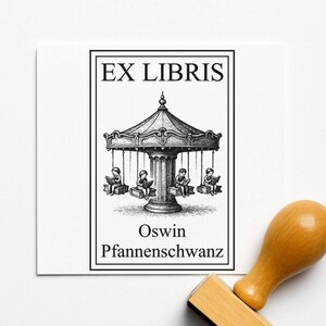Op de afbeelding: Een zwart-wit stempel met de tekst "EX LIBRIS" boven een carrousel illustratie. De naam "Oswin Pfannenschwanz" staat eronder. Een houten stempelhandvat is rechtsonder zichtbaar.
