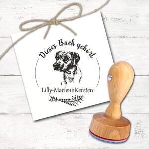 Puede incluir: Un sello de madera con una ilustración de perro y el texto "Dieses Buch gehört" y "Lilly-Marlene Kersten" en tinta negra. El sello tiene un mango de madera y una almohadilla de tinta roja y azul. Un lazo de cuerda natural está atado en la parte superior.