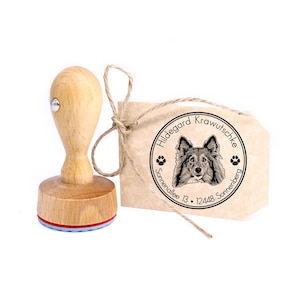 Op de afbeelding: Houten zelf-inktende stempel met een bruine handgreep en een blauwe en rode basis. De stempel heeft een zwart-witte illustratie van een hond met de tekst "Hildegard Krawutschke Sonnenallee 13 · 12448 Sonnenberg" in een cirkel.