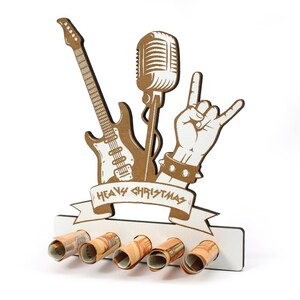 May include: A wooden Christmas decoration featuring a guitar, microphone, and a hand making the rock-and-roll sign. The words "Heavy Christmas" are on a banner. Five rolled-up banknotes are inserted into the base. The design is primarily brown and white.