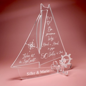 Op de afbeelding: Transparante acryl zeilbootdecoratie met geëtste details. Voorzien van een kompasroos, harten en tekst in script. Inclusief de namen "Silke & Mario" en het getal "2025". De zeilboot staat op een roze achtergrond.
